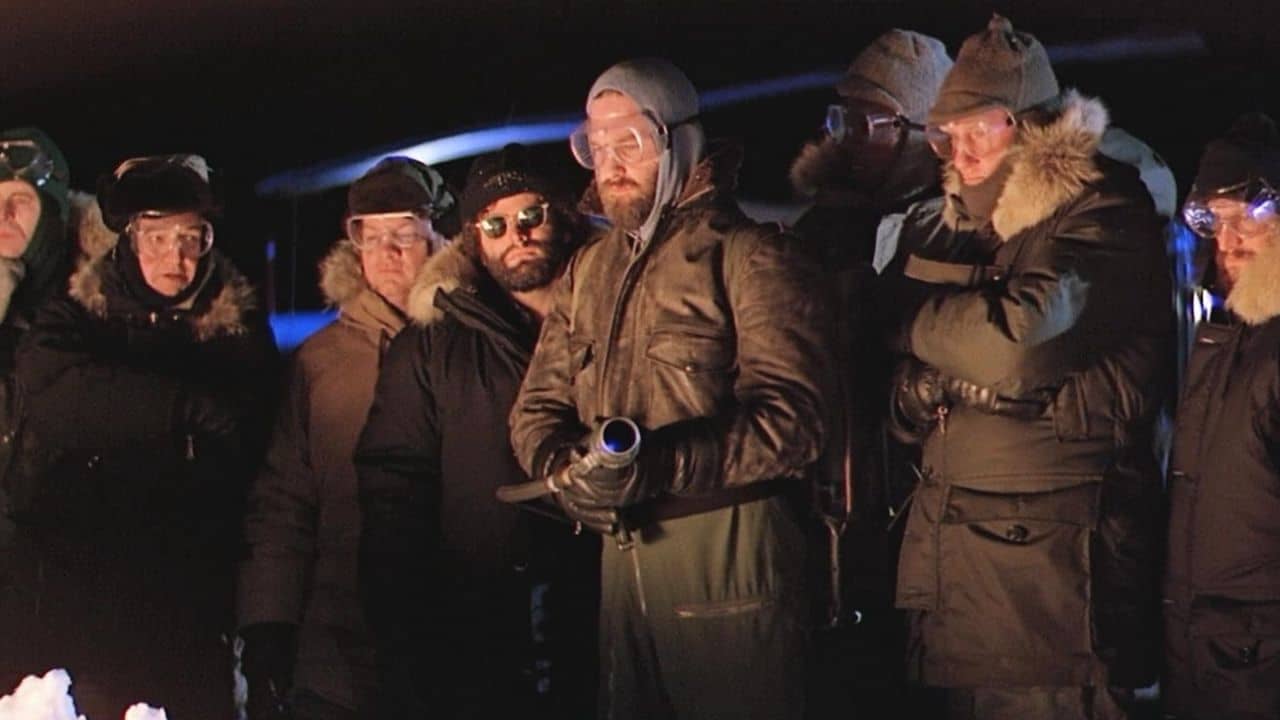 The cast of The Thing look on as R. J. MacReady keeps a flamethrower nearby.