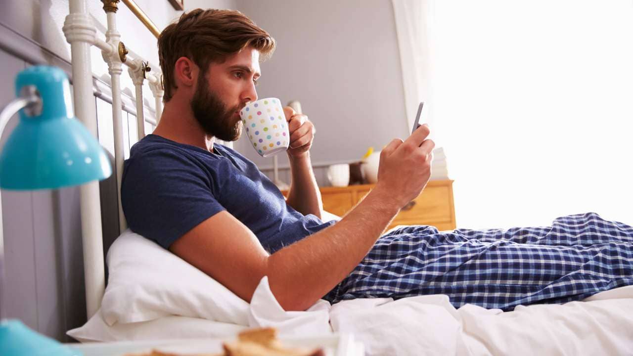 A man at a hotel uses his cell phone in bed while sipping coffee. Breakfast sits next to him on the nightstand.
