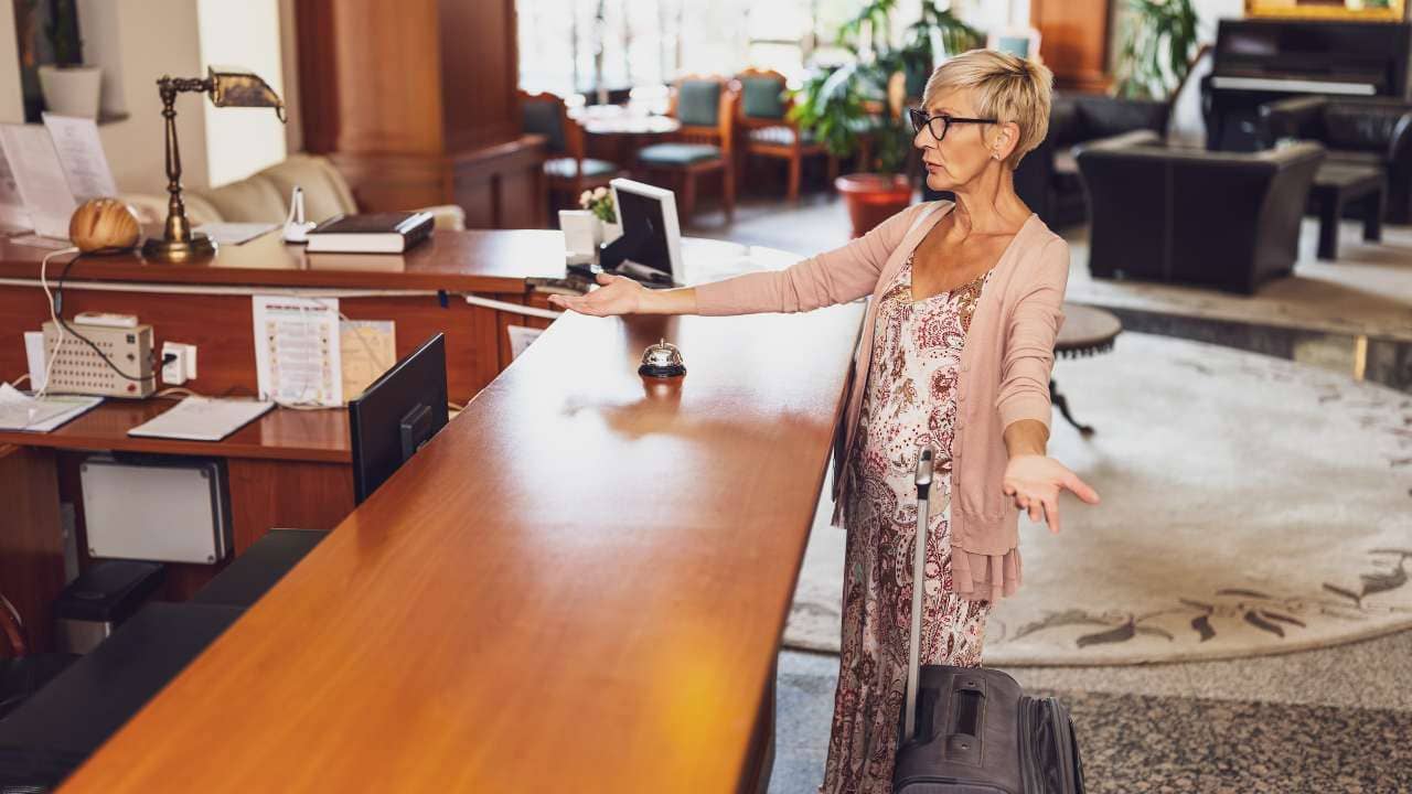 A woman at the front of the hotel reception desk with her luggage holding out her arms, expressing distaste or frustration.