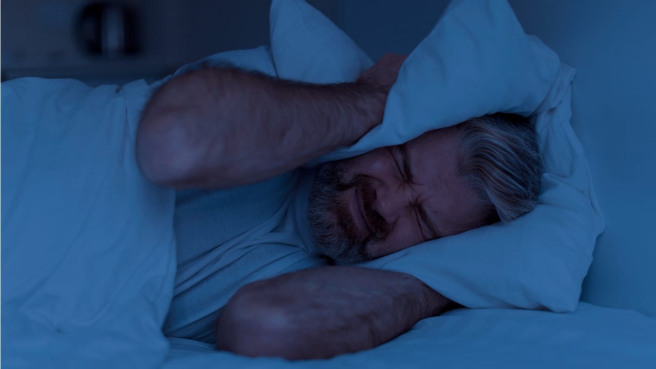 Closeup of angry mature man lying in bed and covering head ears with pillow at night, cannot sleep, suffering from noisy neighbours or snoring spouse partner