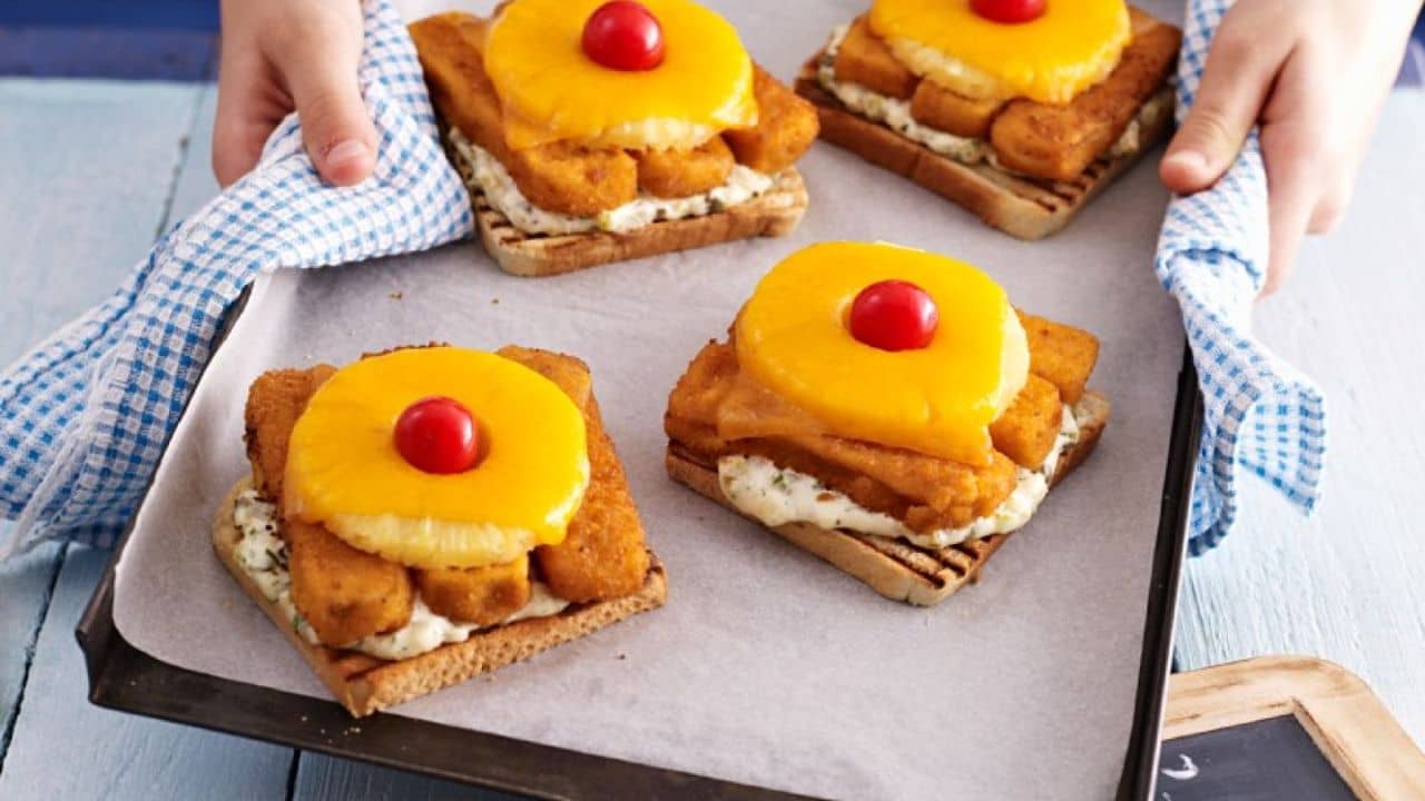 A woman holding out a baking pan with multiple pieces of bread, topped with fish sticks, pineapple and plated as if it were a pineapple upside down cake.
