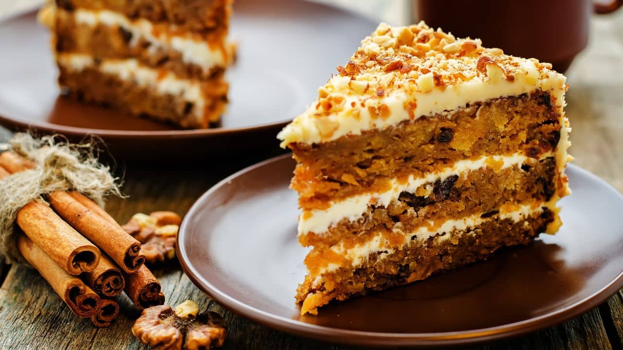 A beautifully crafted slice of carrot cake placed on a brown plate, next to a second slice in the same way in the background - they both rest next to displayed cinnamon sticks. 