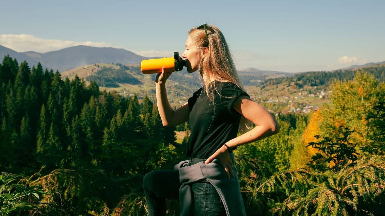 A woman drinks from a reusable water bottle while exploring outdoors.