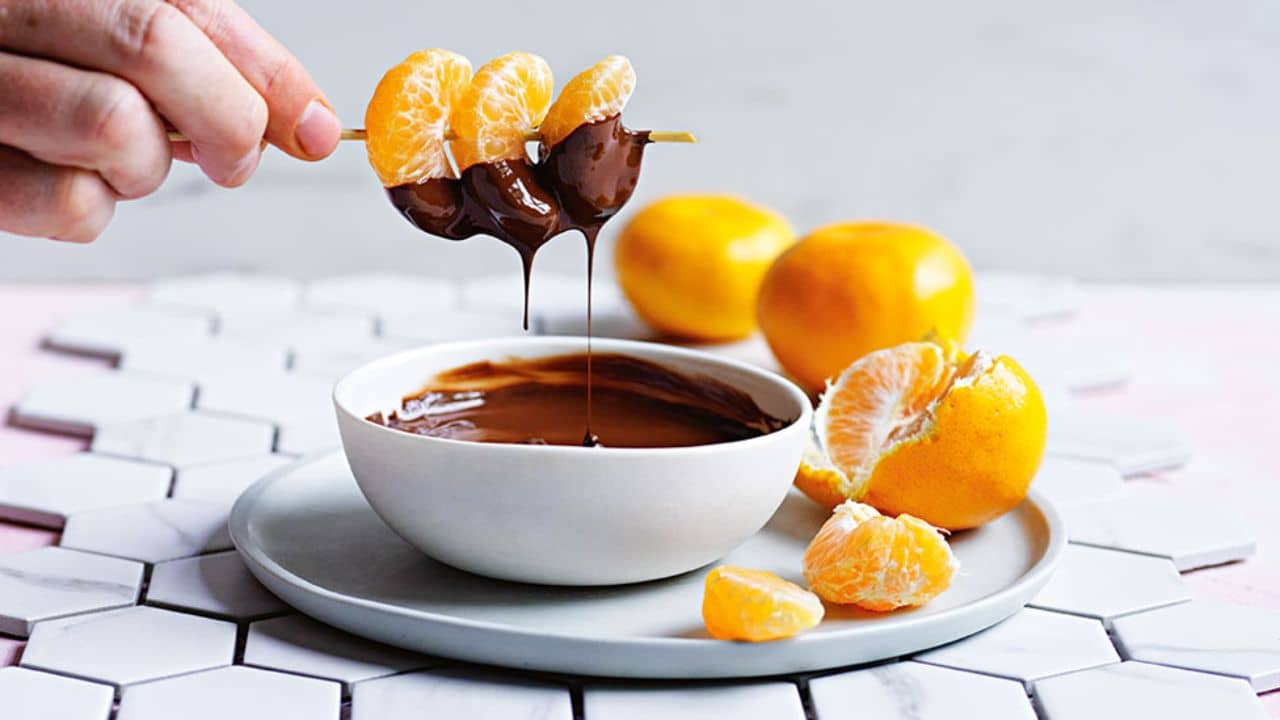 A caucasian hand dipping multiple small mandarin orange slices on a skewer into chocolate fondue. 